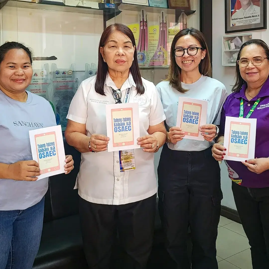 Four women stand in a Barangay office holding booklets, smiling. Trophy-filled cabinet and framed picture in the background convey achievement.
