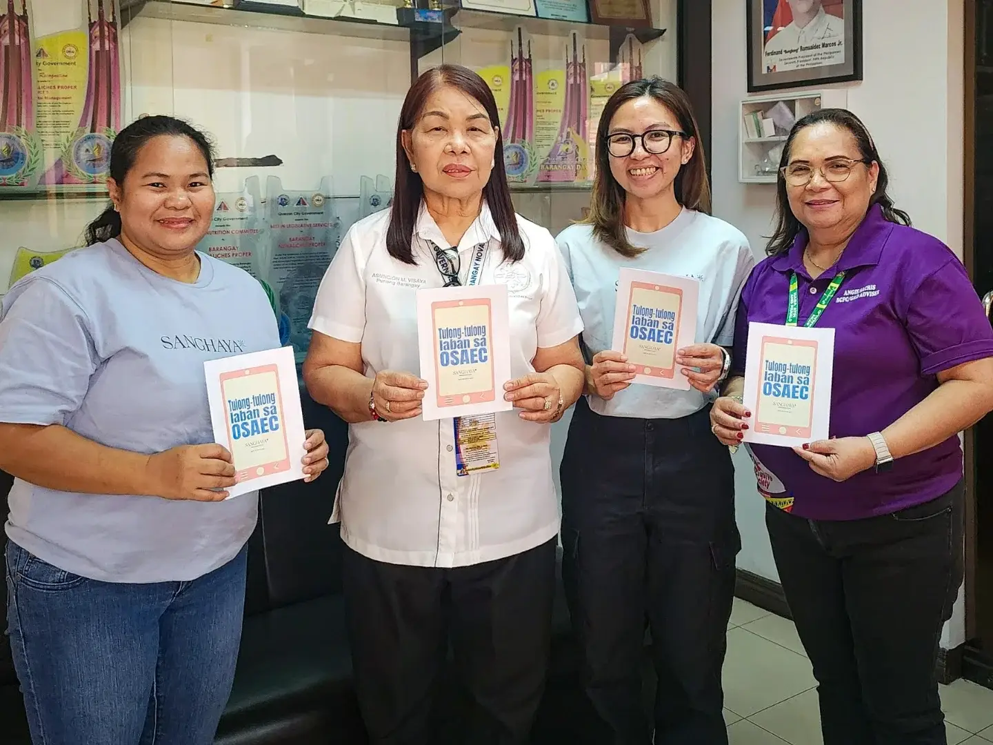Four women stand in a Barangay office holding booklets, smiling. Trophy-filled cabinet and framed picture in the background convey achievement.
