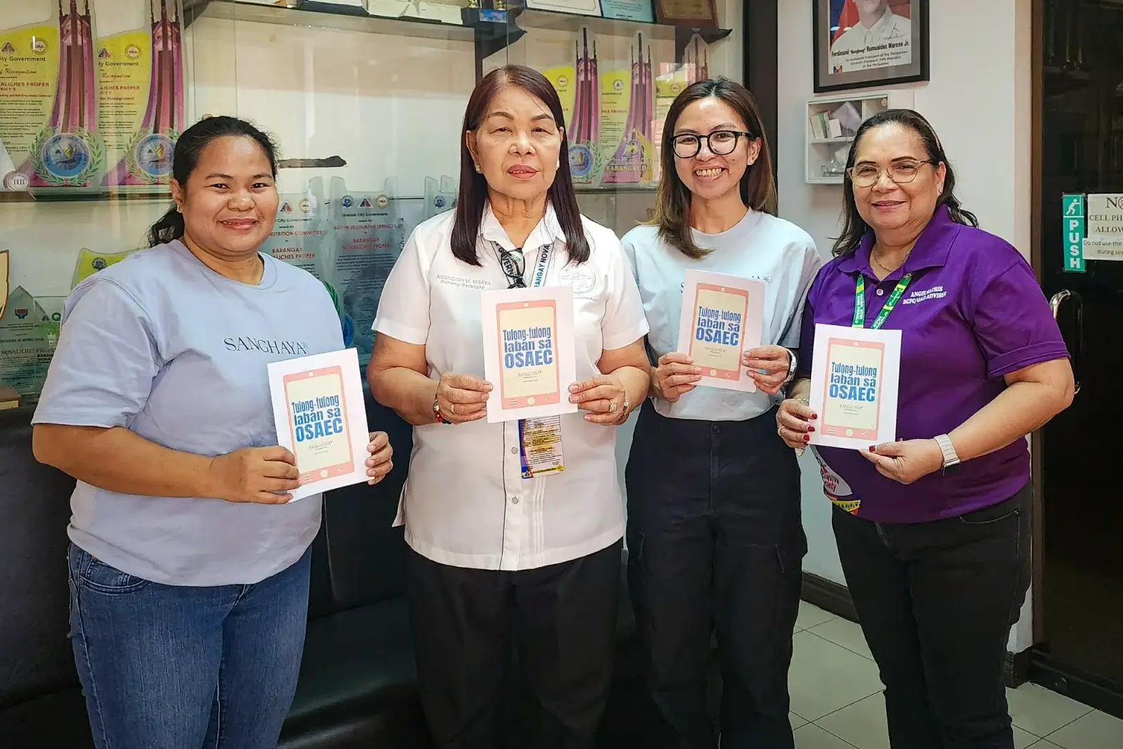 Four women stand in a Barangay office holding booklets, smiling. Trophy-filled cabinet and framed picture in the background convey achievement.
