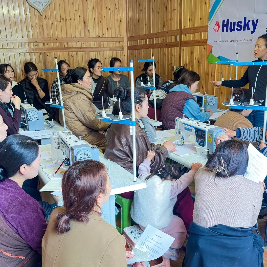 A group of women in a wooden room are attentively learning sewing skills. A teacher stands in front of sewing machines, under a banner thanking sponsors.