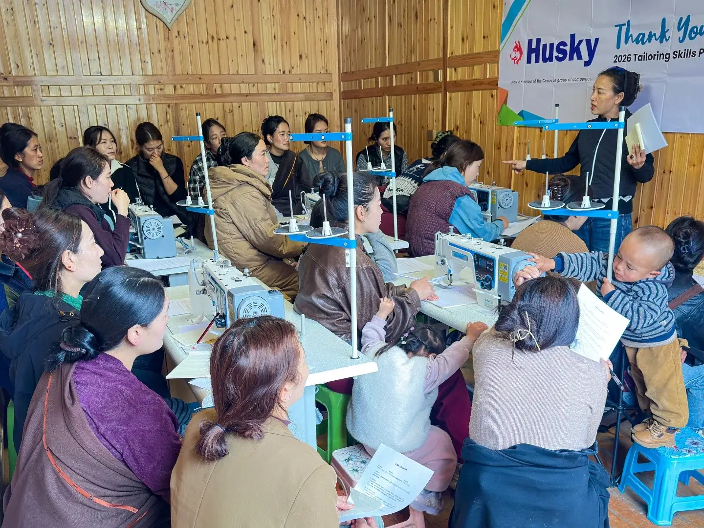 A group of women in a wooden room are attentively learning sewing skills. A teacher stands in front of sewing machines, under a banner thanking sponsors.