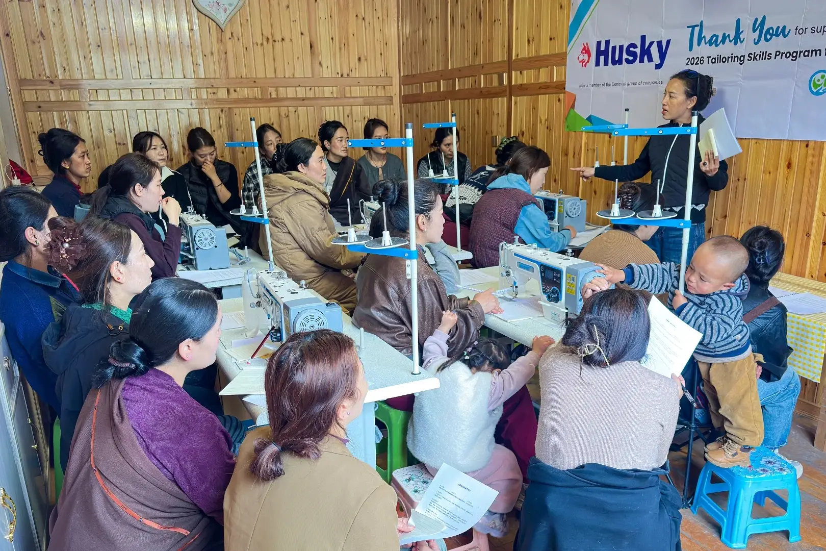 A group of women in a wooden room are attentively learning sewing skills. A teacher stands in front of sewing machines, under a banner thanking sponsors.