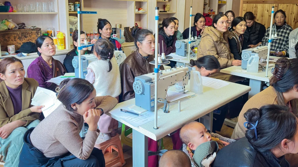 A group of women attentively sit in a sewing class, surrounded by sewing machines. The room conveys focus and community learning. A baby is seen in the foreground.