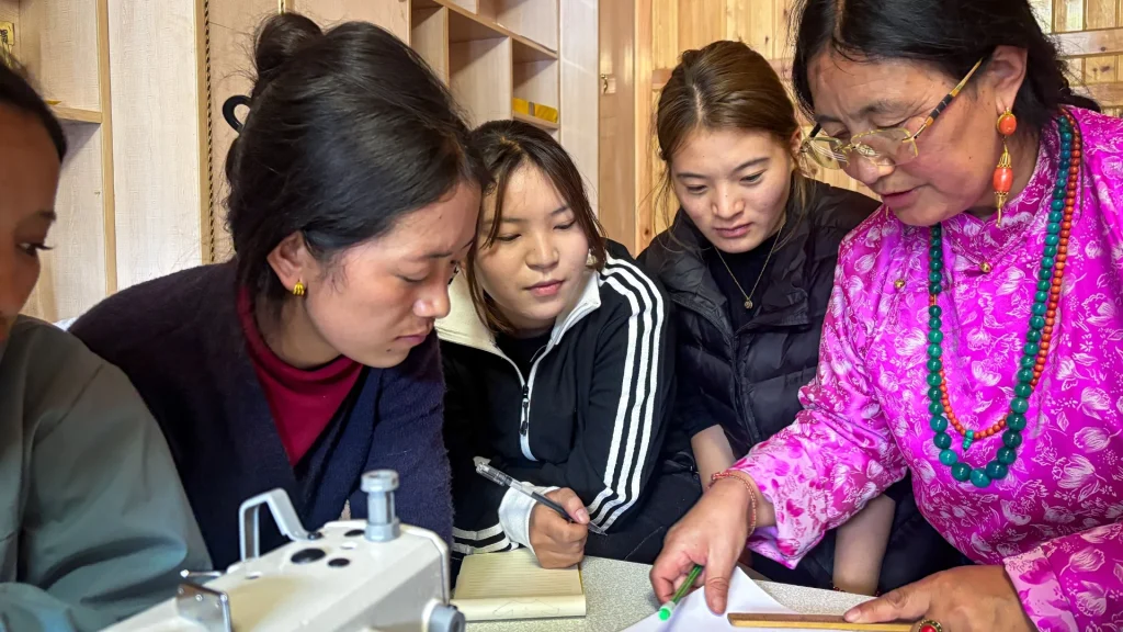 A group of women intently observe a woman in a pink dress pointing to a paper near a sewing machine. They appear focused and engaged.A group of women intently observe a woman in a pink dress pointing to a paper near a sewing machine. They appear focused and engaged.