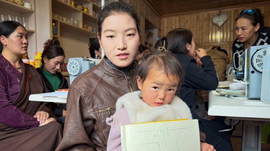 A woman in a leather jacket holds a child with a drawing. They sit in a cozy room with sewing machines and other women engaged in conversation.