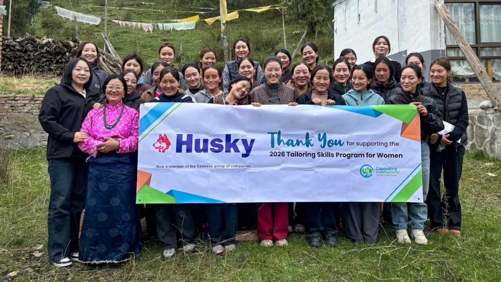 A group of women smiling and holding a banner that reads, "Husky: Thank You for supporting the 2026 Tailoring Skills Program for Women." Outdoor setting with greenery.