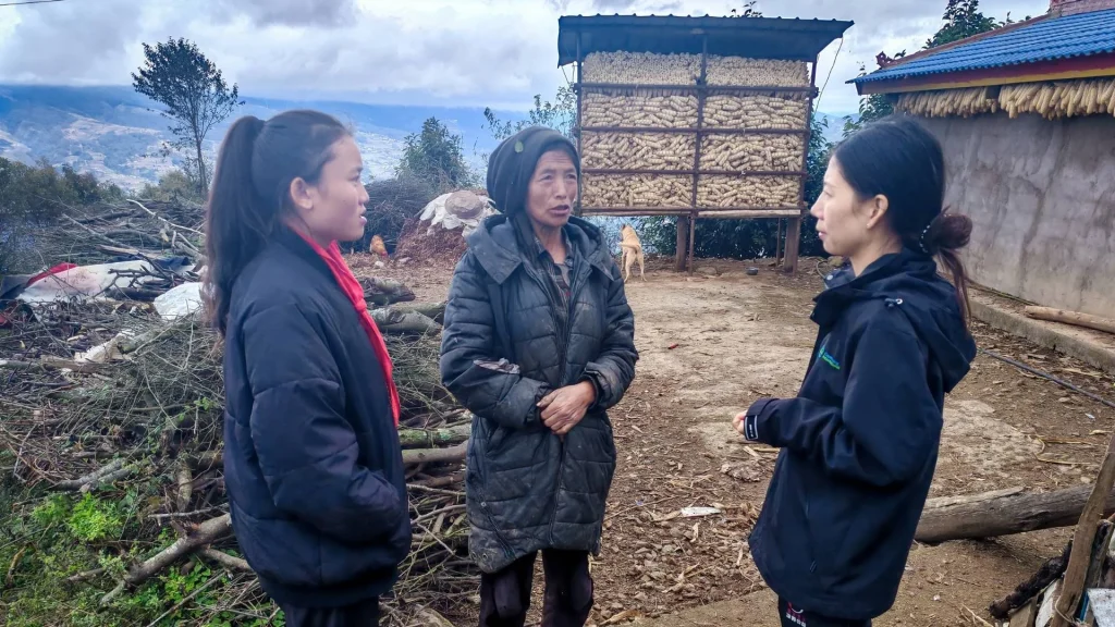 Three women stand outdoors in conversation on a dirt path. One woman wears a hooded coat, and the other two are in jackets. Behind them is a large stacked pile of corn and a blue-roofed structure. The background features trees and a mountain view, creating a rural setting. The mood appears serious and focused.