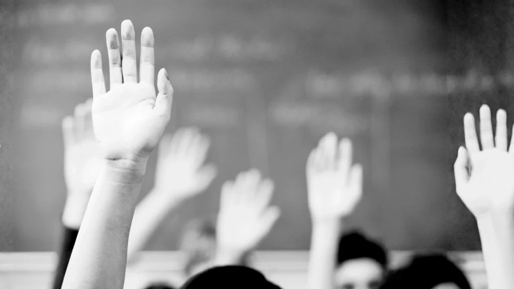 Black and white image of multiple hands raised in front of a blurred chalkboard. The scene suggests an active classroom setting with student engagement.