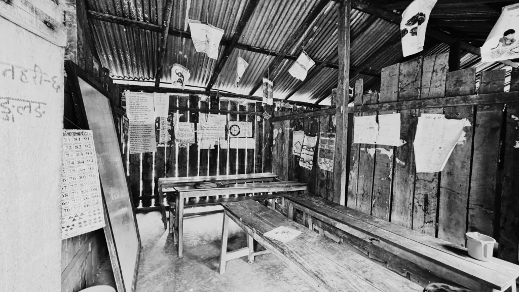 Black and white image of a simple, rustic classroom with wooden benches and walls. Posters and charts are tacked to the walls, creating an educational atmosphere.