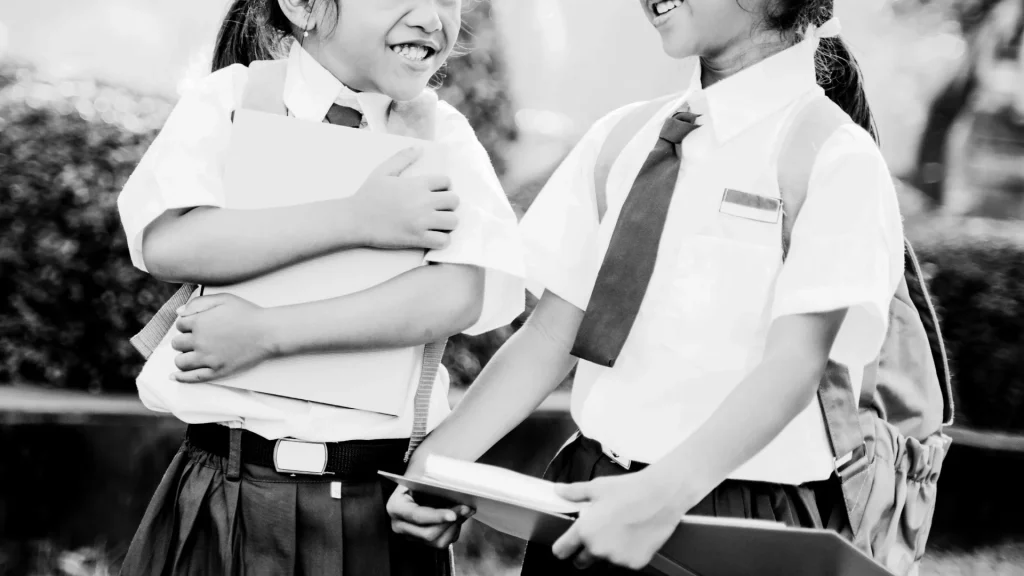 Black and white image of two laughing schoolgirls in uniform, holding books and backpacks, suggesting a joyful and friendly moment in a schoolyard.