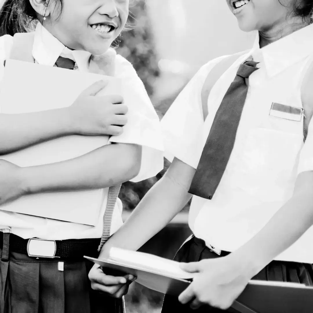 Black and white image of two laughing schoolgirls in uniform, holding books and backpacks, suggesting a joyful and friendly moment in a schoolyard.
