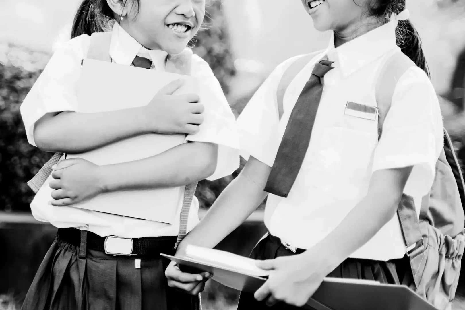 Black and white image of two laughing schoolgirls in uniform, holding books and backpacks, suggesting a joyful and friendly moment in a schoolyard.