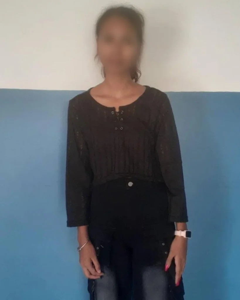 A person stands against a two-tone wall, wearing a black top and jeans. They have a bracelet and a watch, with relaxed body language. Their face is blurred for privacy.