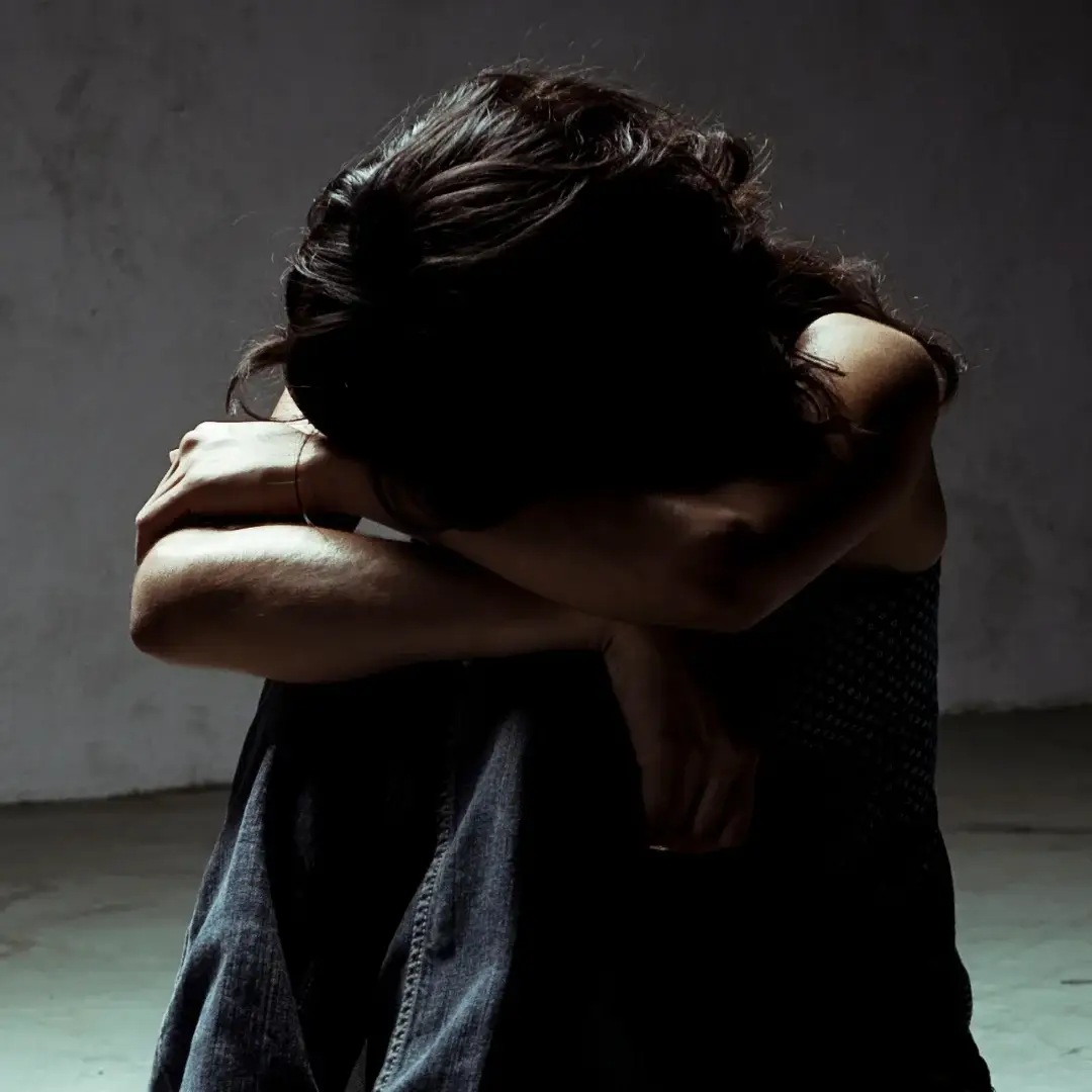 A person sits on a floor in a dim room, head buried in crossed arms, conveying sadness or isolation. The scene evokes a somber, reflective mood.