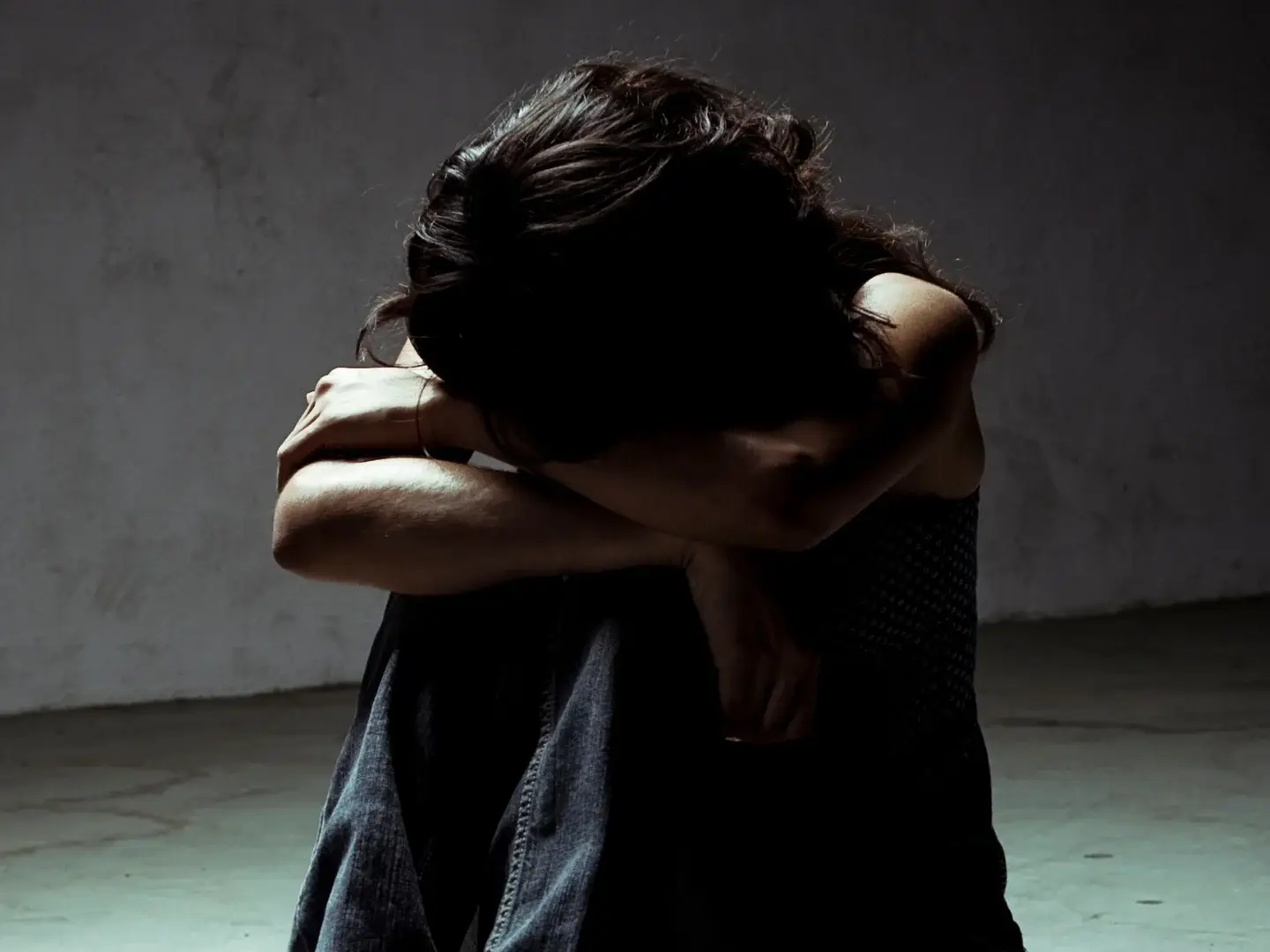 A person sits on a floor in a dim room, head buried in crossed arms, conveying sadness or isolation. The scene evokes a somber, reflective mood.