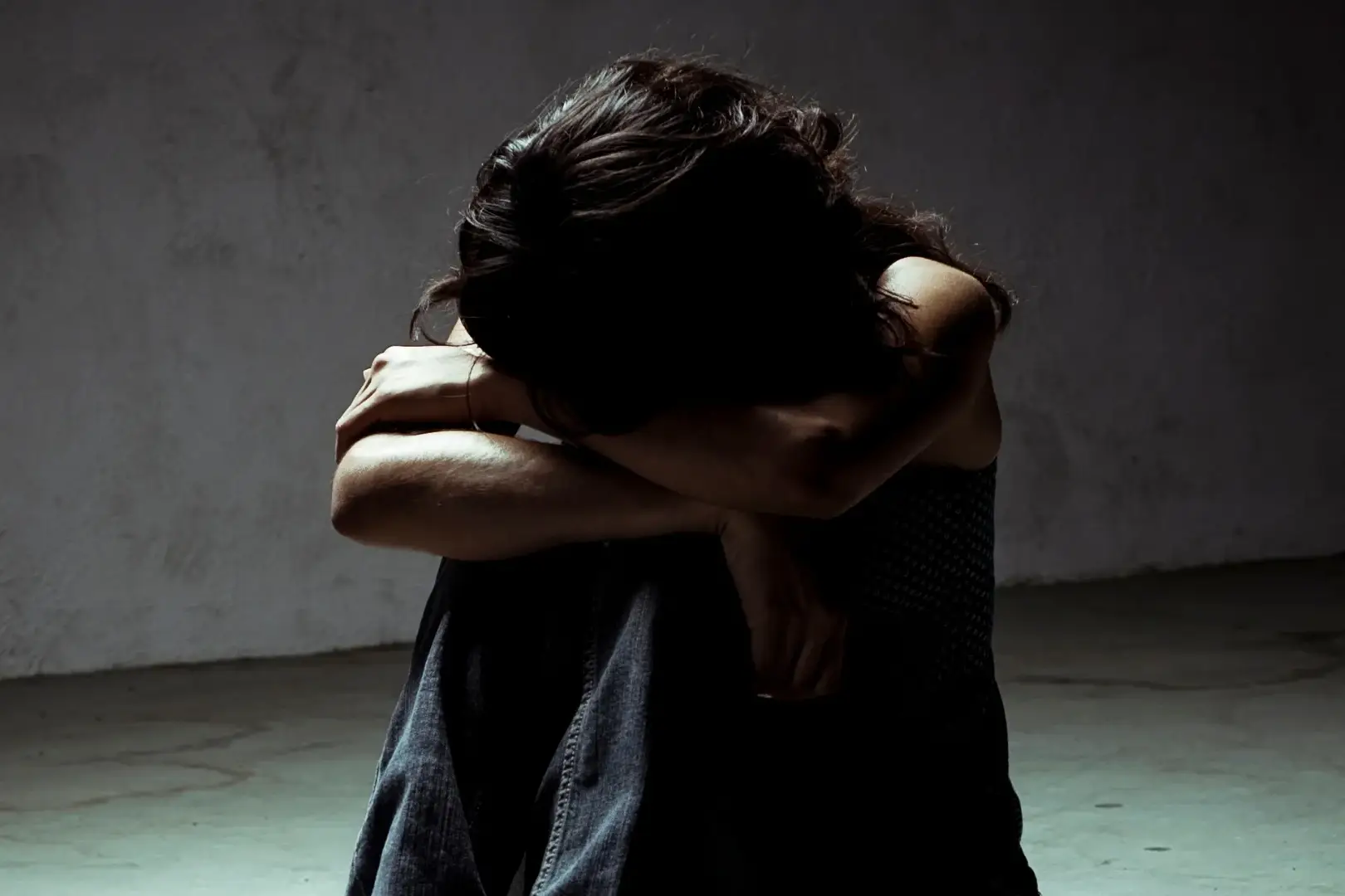 A person sits on a floor in a dim room, head buried in crossed arms, conveying sadness or isolation. The scene evokes a somber, reflective mood.