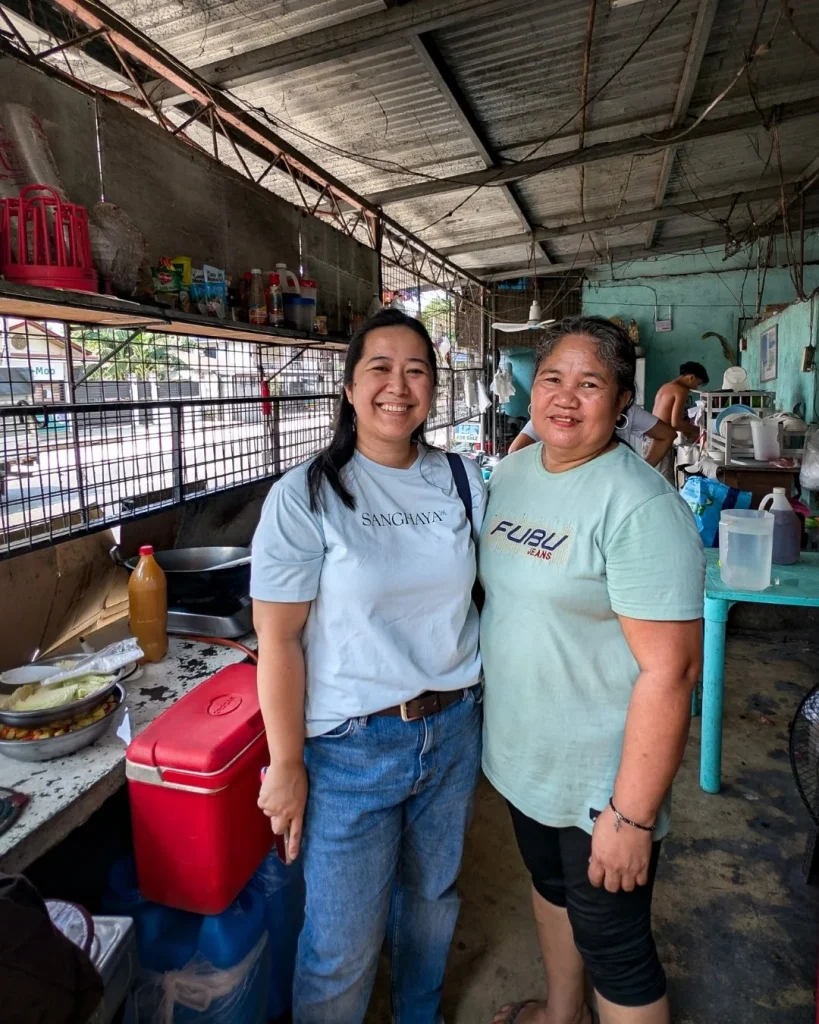 Two women are smiling in a rustic kitchen with a metal roof. Various kitchen supplies and a red cooler are on the counter. The atmosphere is warm and friendly.