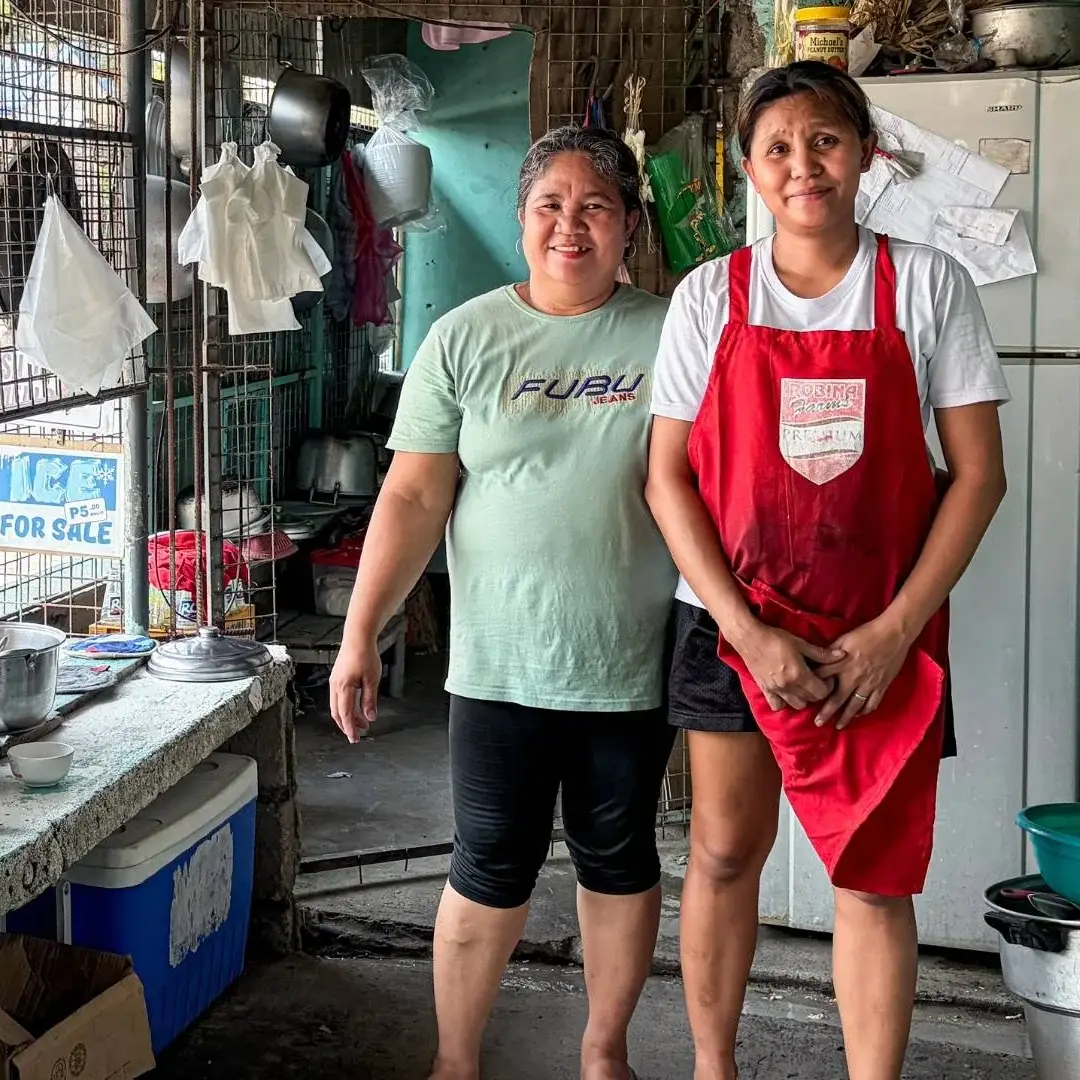 Two women stand in a rustic kitchen, one wearing an apron and the other smiling. Pots and kitchen items surround them, conveying a warm, homely atmosphere.