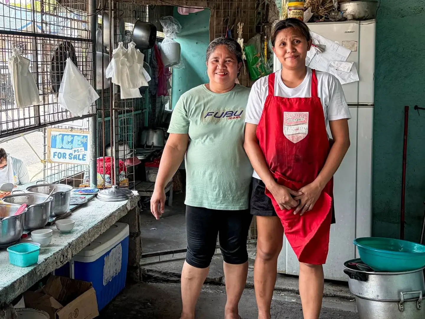 Two women stand in a rustic kitchen, one wearing an apron and the other smiling. Pots and kitchen items surround them, conveying a warm, homely atmosphere.