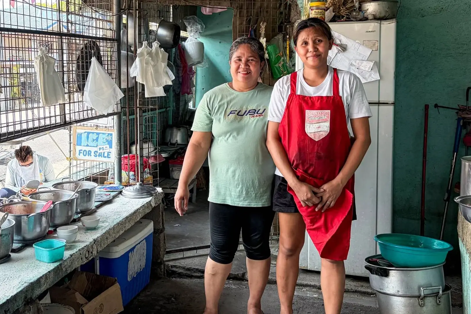 Two women stand in a rustic kitchen, one wearing an apron and the other smiling. Pots and kitchen items surround them, conveying a warm, homely atmosphere.
