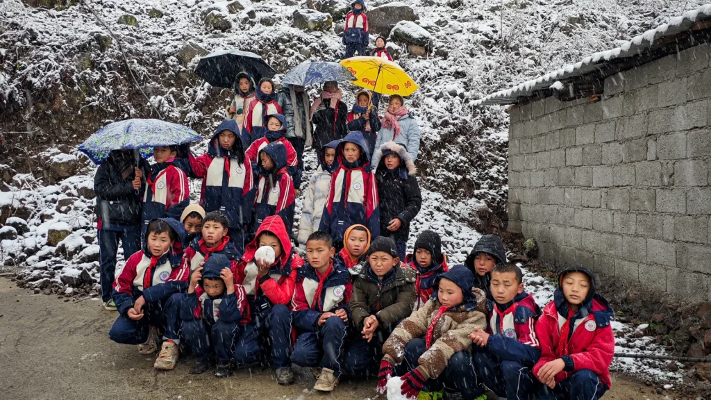 A group of children in red jackets and winter clothes gather outdoors amidst gentle snowfall, holding colorful umbrellas. The scene is cheerful and lively.