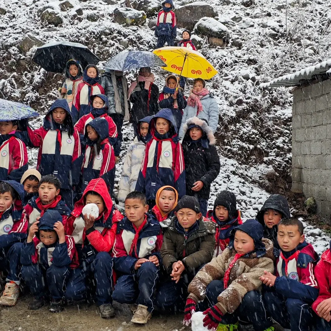 A group of children in red jackets and winter clothes gather outdoors amidst gentle snowfall, holding colorful umbrellas. The scene is cheerful and lively.