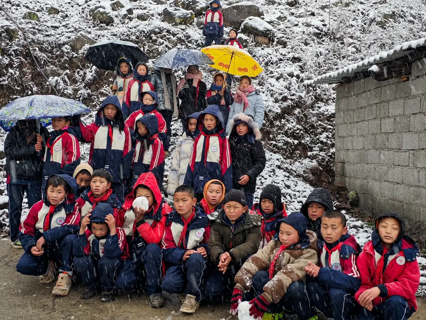 A group of children in red jackets and winter clothes gather outdoors amidst gentle snowfall, holding colorful umbrellas. The scene is cheerful and lively.