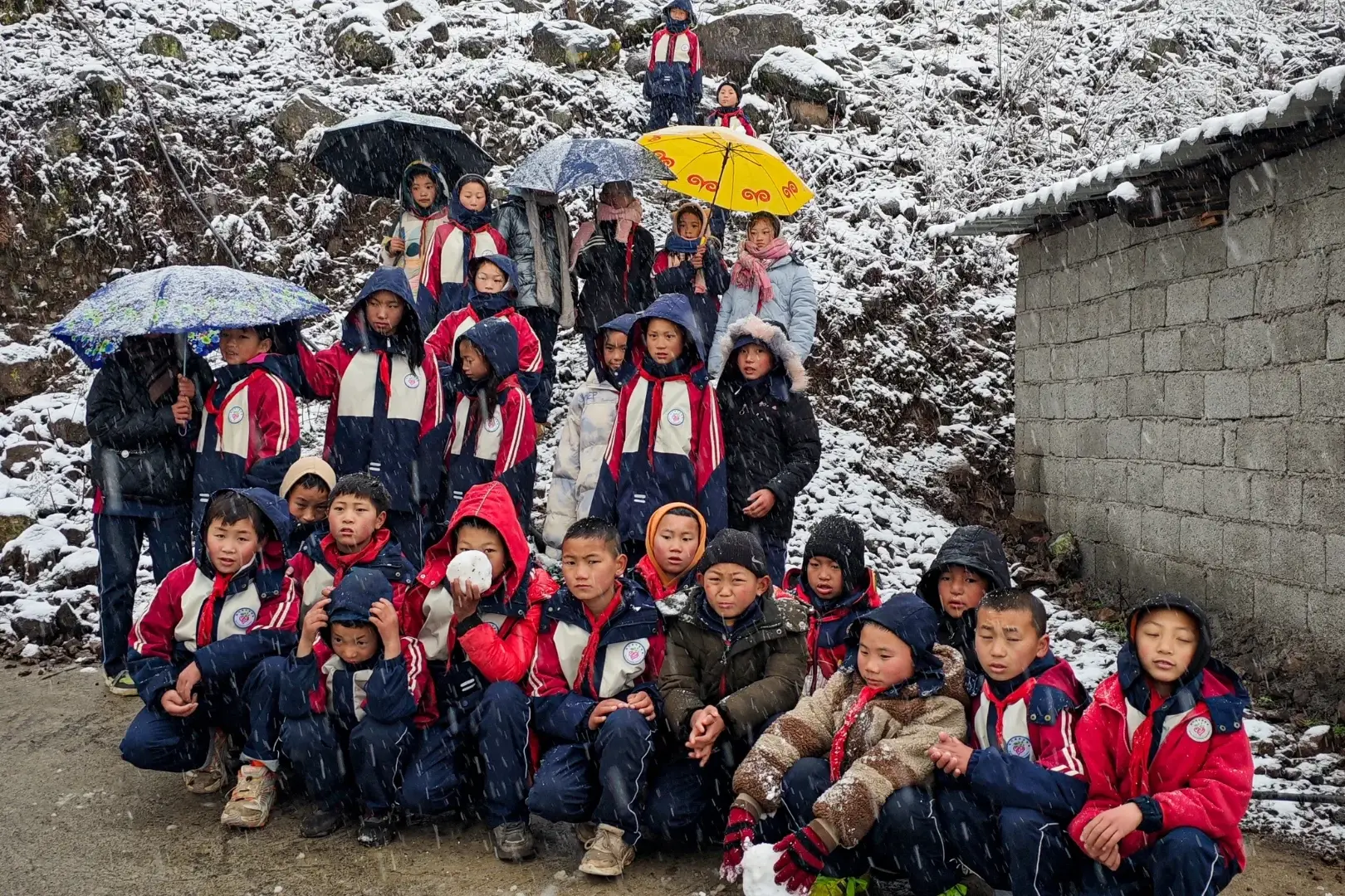 A group of children in red jackets and winter clothes gather outdoors amidst gentle snowfall, holding colorful umbrellas. The scene is cheerful and lively.