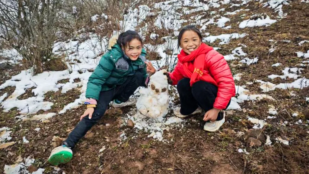 Two smiling children in winter jackets, one green and one red, crouch beside a small snowman. Snow patches are on the ground, and bare bushes surround them.