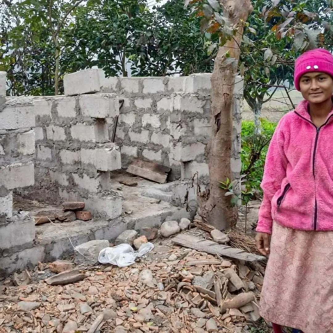 A woman in a pink jacket and hat stands beside an incomplete brick structure. The scene is outdoors with trees and scattered building materials, evoking a sense of resilience and construction progress.