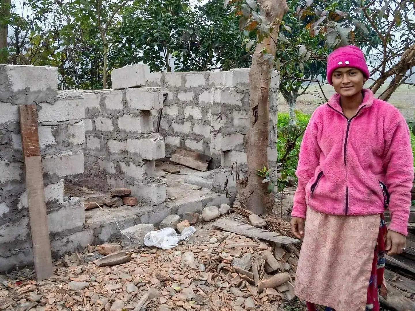A woman in a pink jacket and hat stands beside an incomplete brick structure. The scene is outdoors with trees and scattered building materials, evoking a sense of resilience and construction progress.