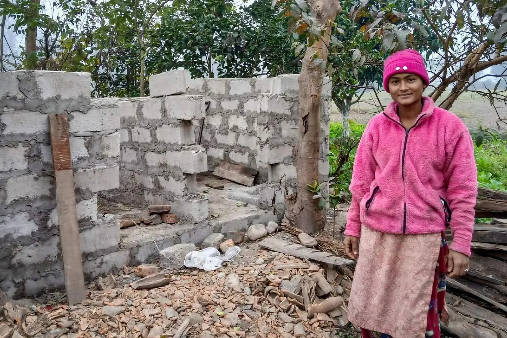 A woman in a pink jacket and hat stands beside an incomplete brick structure. The scene is outdoors with trees and scattered building materials, evoking a sense of resilience and construction progress.