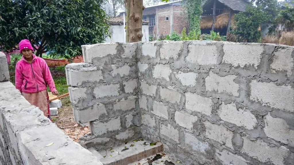 A woman in a pink jacket and hat stands beside an incomplete brick structure. The scene is outdoors with trees and scattered building materials, evoking a sense of resilience and construction progress.