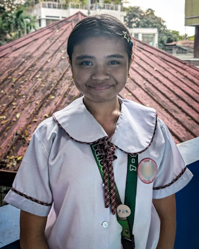 A smiling girl in a school uniform with a plaid tie and collar stands in front of a rust-colored roof. The background includes trees and buildings.