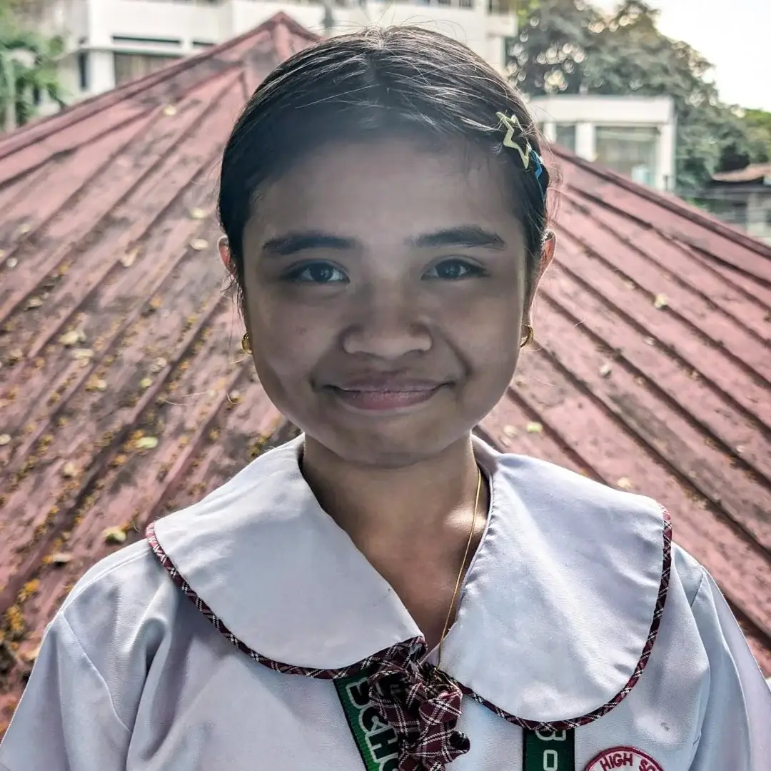 A smiling girl in a school uniform with a plaid tie and collar stands in front of a rust-colored roof. The background includes trees and buildings.