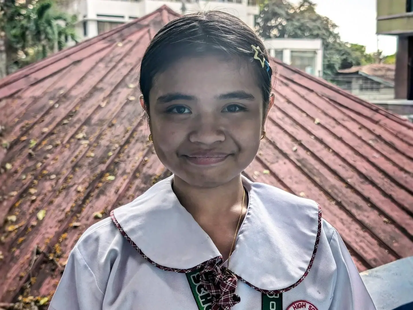 A smiling girl in a school uniform with a plaid tie and collar stands in front of a rust-colored roof. The background includes trees and buildings.
