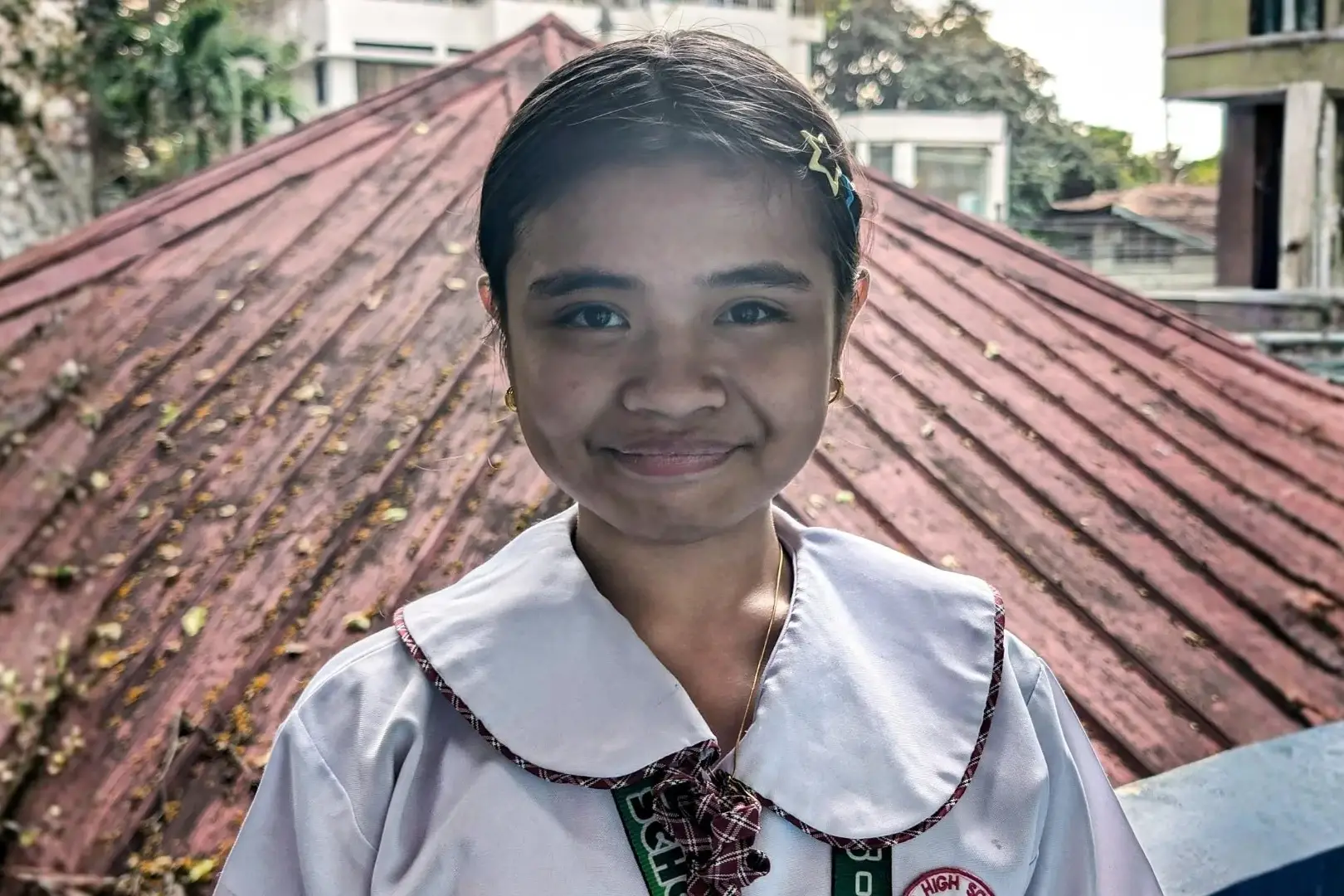 A smiling girl in a school uniform with a plaid tie and collar stands in front of a rust-colored roof. The background includes trees and buildings.