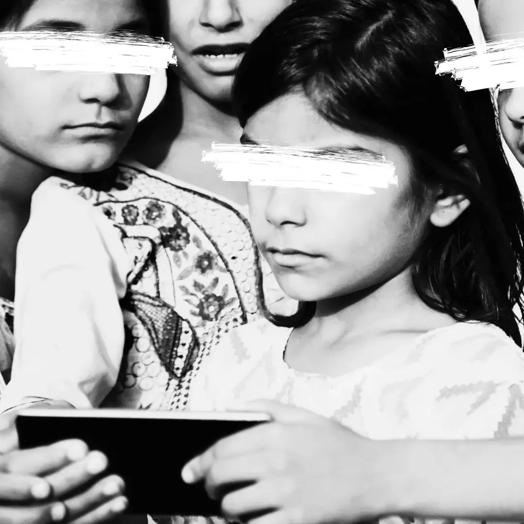 Four children, eyes obscured, huddle closely, intrigued by a smartphone. They wear embroidered outfits, the mood is focused and curious. Black and white.