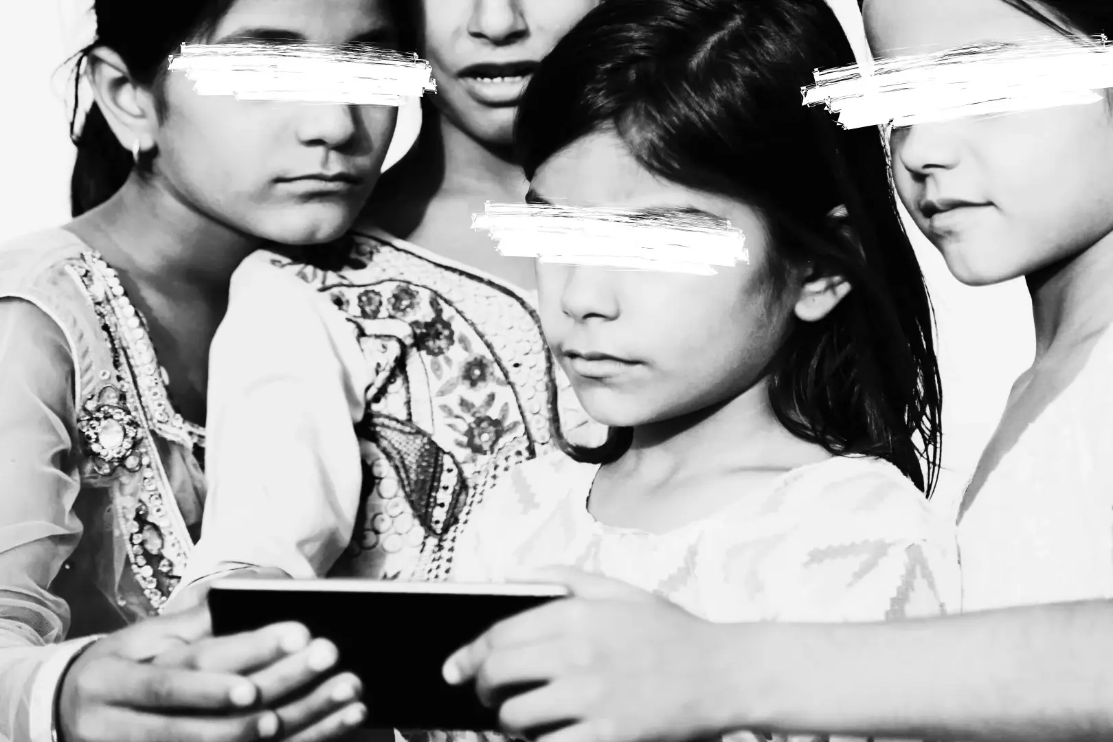 Four children, eyes obscured, huddle closely, intrigued by a smartphone. They wear embroidered outfits, the mood is focused and curious. Black and white.
