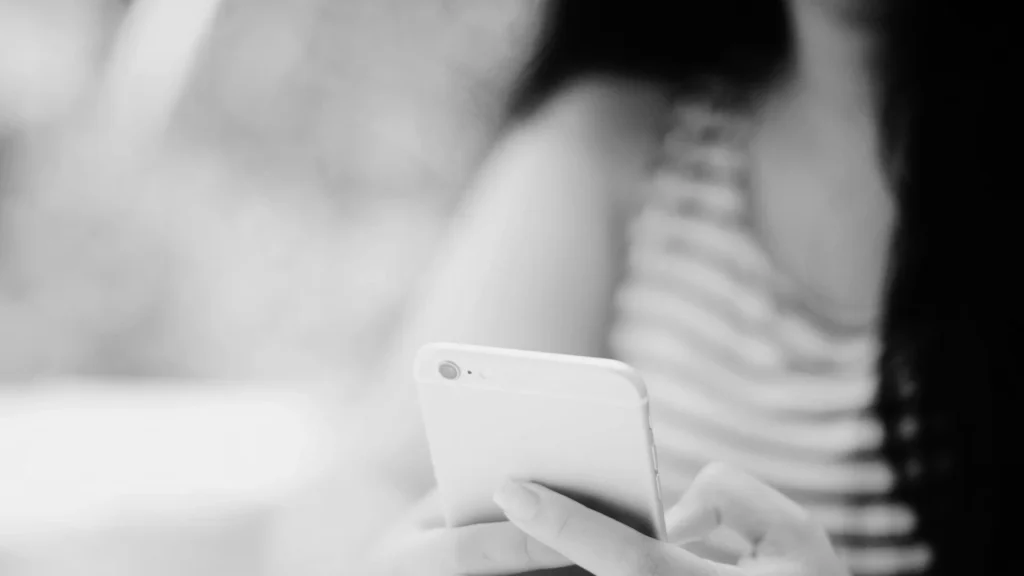 Black and white image of a person holding a smartphone, wearing a striped tank top. The atmosphere is calm, focusing on the use of technology.