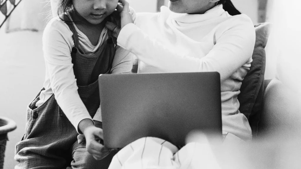 A black-and-white image shows a woman and a child sitting together on a couch, looking at a laptop. The atmosphere is warm and focused.