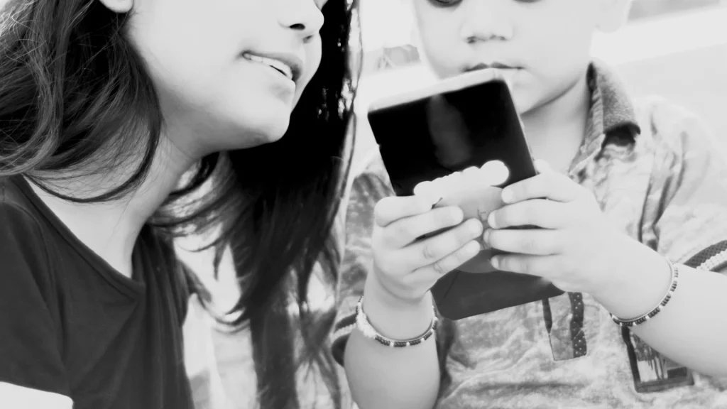A woman and a young child share a moment, focusing on a smartphone the child is holding. The black and white image conveys curiosity and connection.