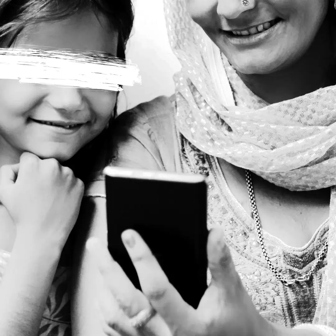 A joyful young girl and a woman in traditional attire smile as they look at a smartphone. The black and white image conveys warmth and connection.