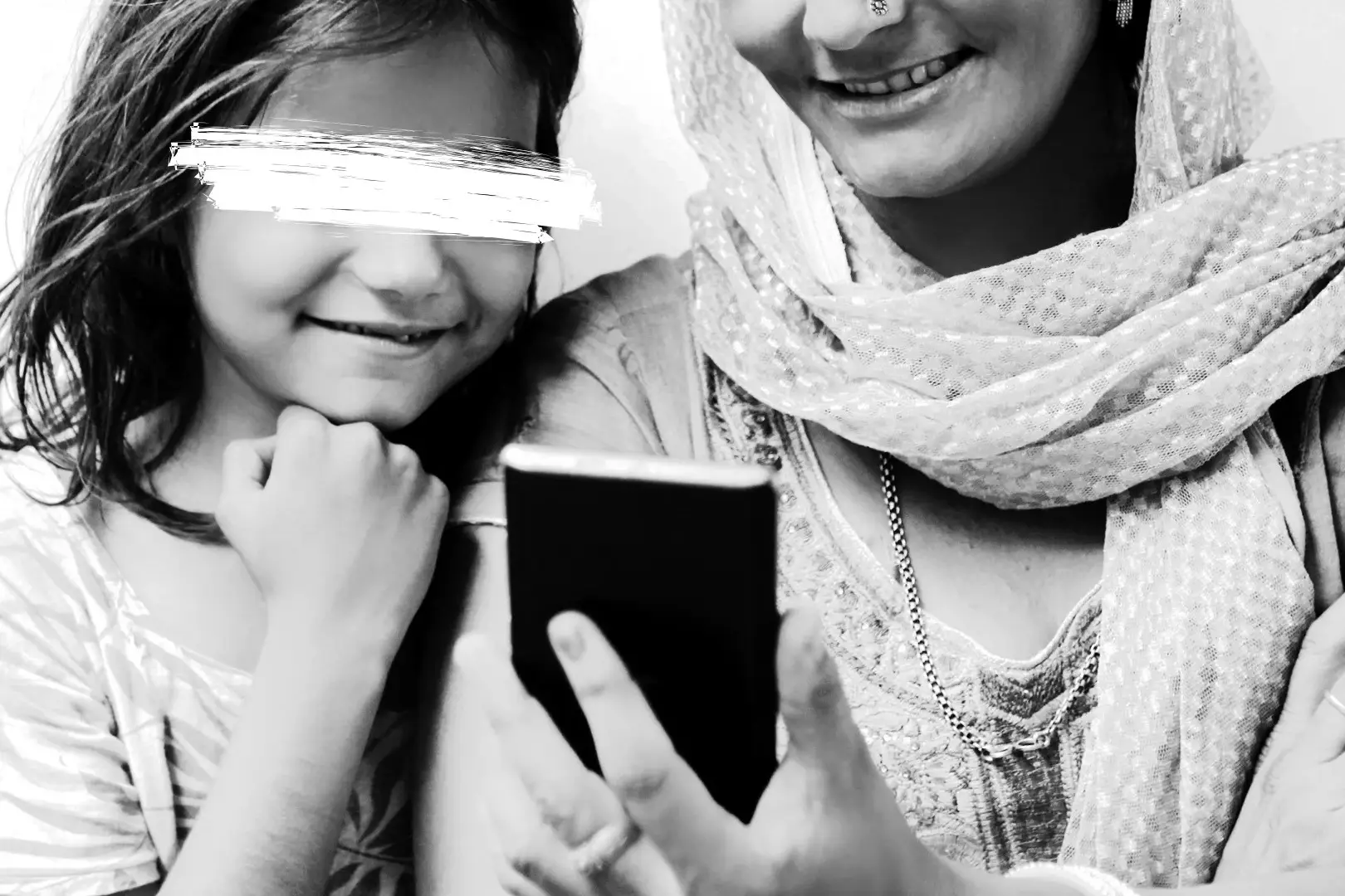 A joyful young girl and a woman in traditional attire smile as they look at a smartphone. The black and white image conveys warmth and connection.