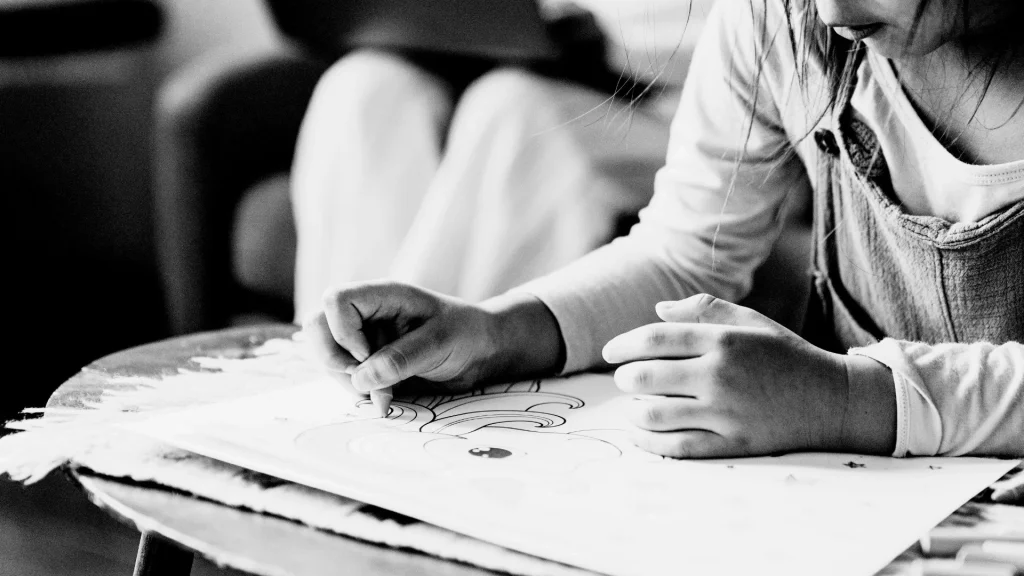 A child focuses intently on coloring a rainbow with a crayon on paper at a small table. The scene is black and white, conveying a calm, creative atmosphere.