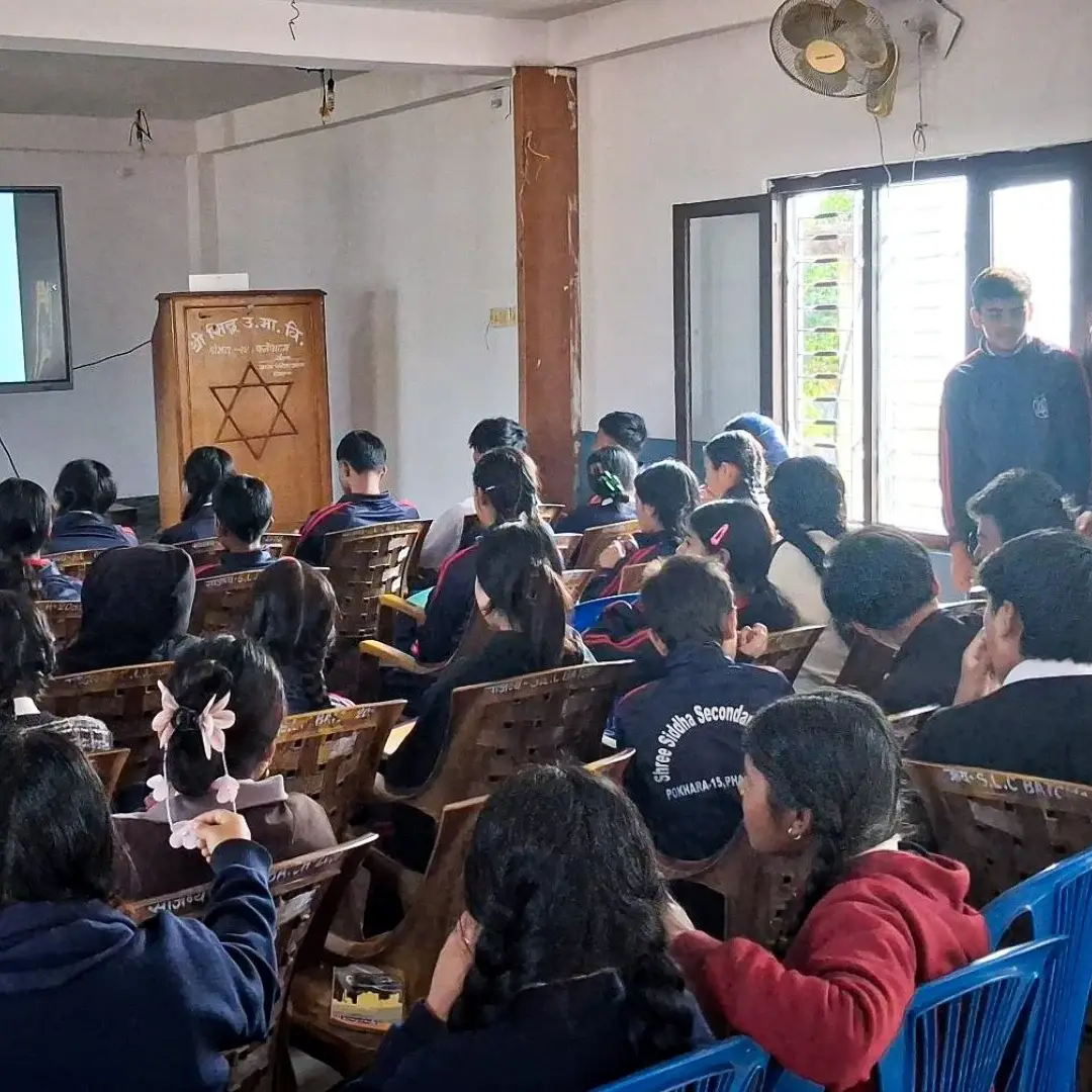A teacher is presenting a lesson to a classroom filled with attentive students seated in rows. A projector screen displays text, creating an educational atmosphere.
