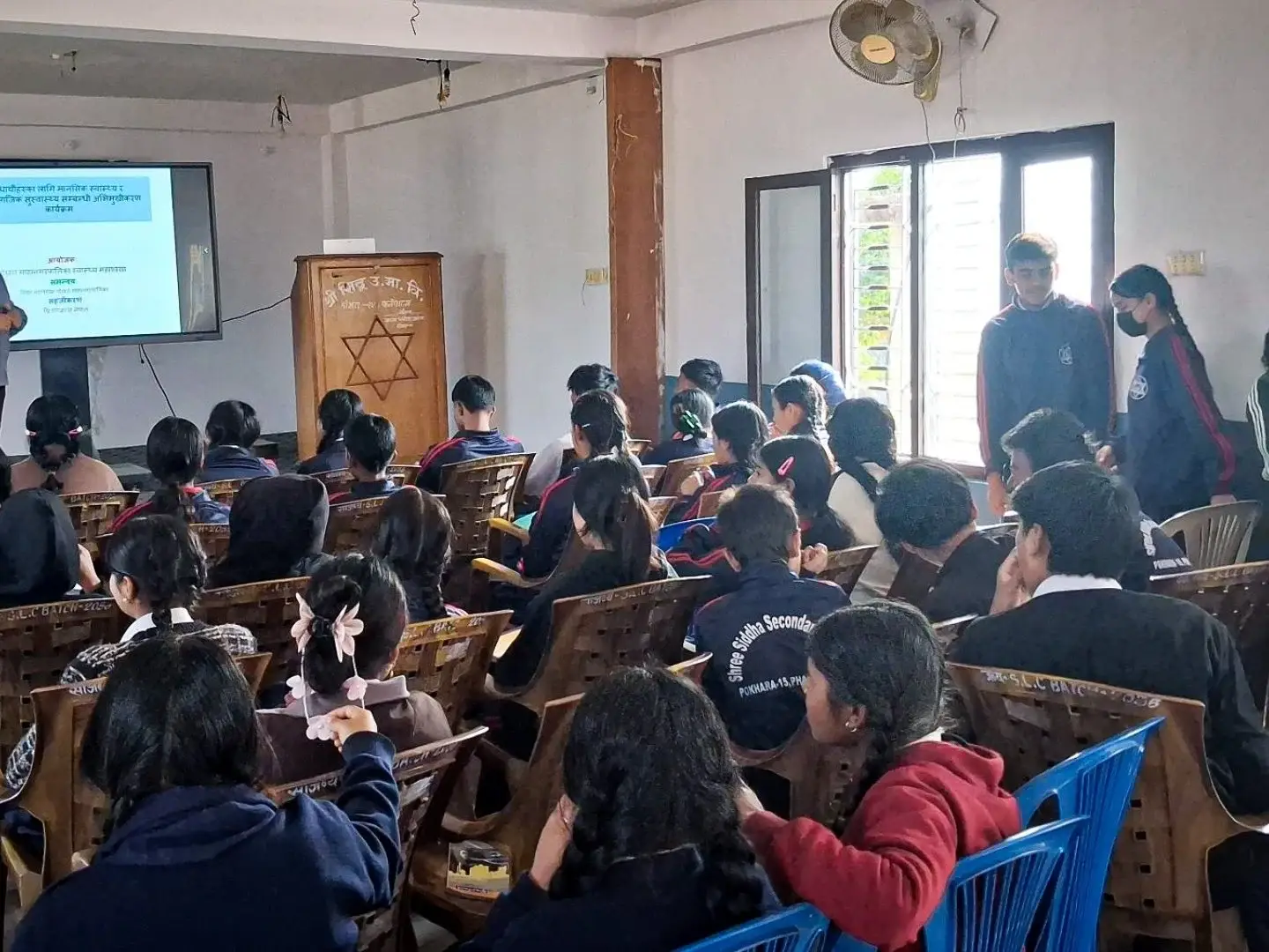 A teacher is presenting a lesson to a classroom filled with attentive students seated in rows. A projector screen displays text, creating an educational atmosphere.