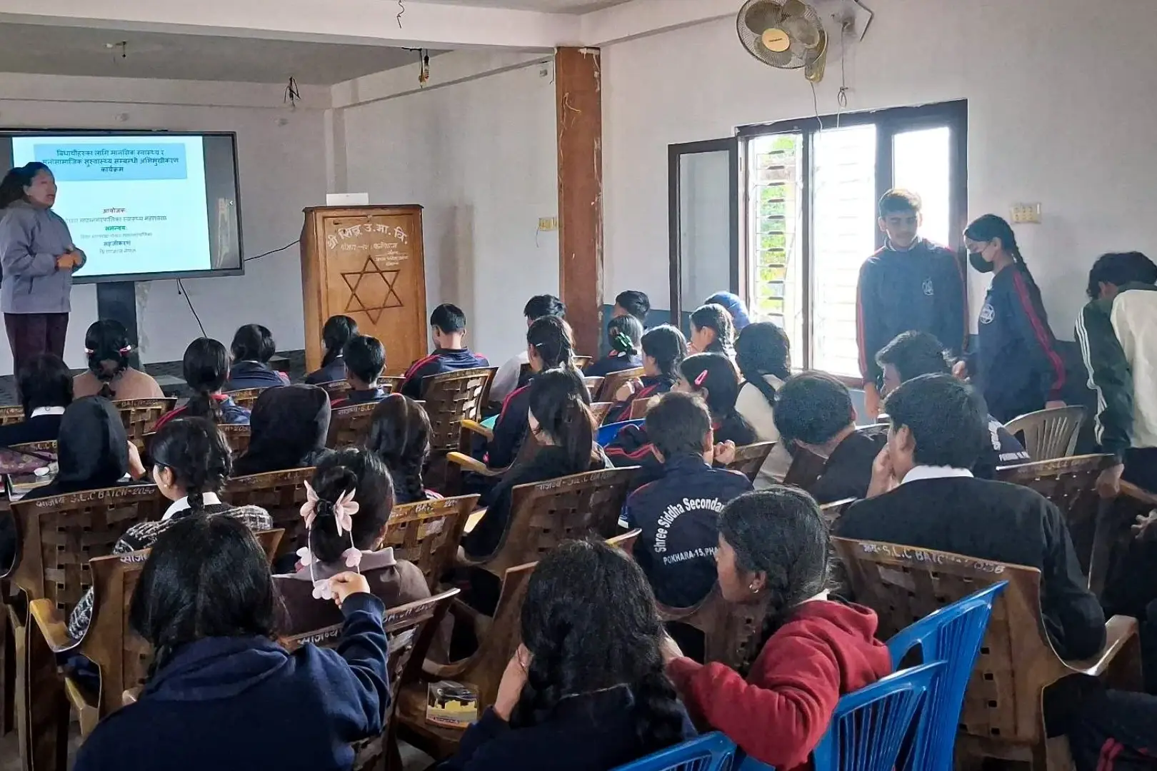 A teacher is presenting a lesson to a classroom filled with attentive students seated in rows. A projector screen displays text, creating an educational atmosphere.