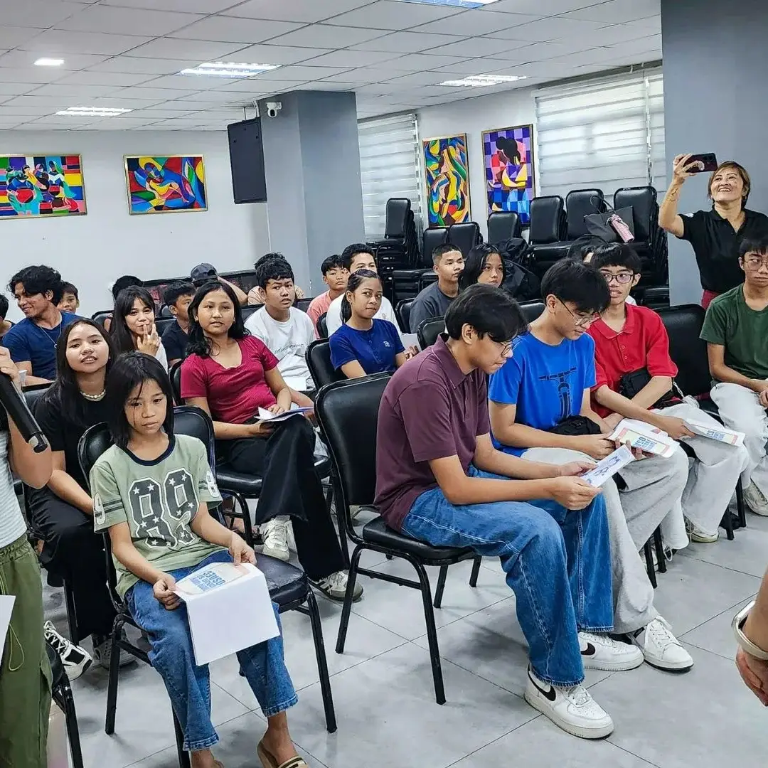 A young girl speaks into a microphone in a classroom, holding a paper. Students sit attentively, some holding papers. A woman in watches.