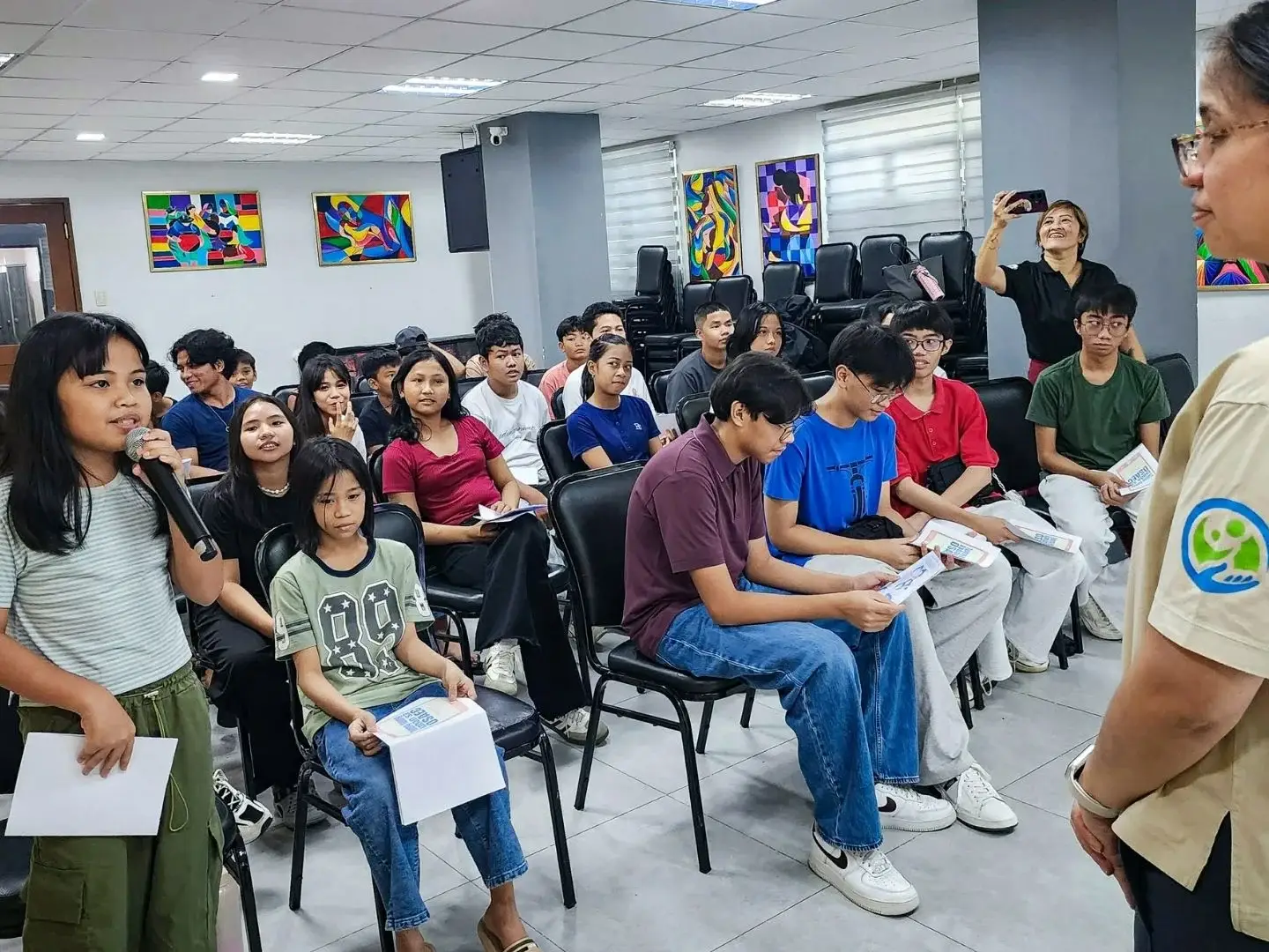 A young girl speaks into a microphone in a classroom, holding a paper. Students sit attentively, some holding papers. A woman in watches.